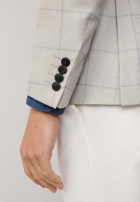 Ręka i nadgarstek w beżowej, kratkowanej marynarce z czterema ciemnymi guzikami, noszące niebieską koszulę i białe spodnie na neutralnym tle.