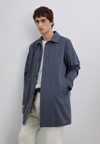 Joven vestido con un abrigo azul abotonado, camisa blanca y pantalones beige, de pie con una mano en el bolsillo frente a un fondo claro y liso.