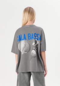 Pilkas oversized marškinėliai su dideliu mėlynu tekstu "CALA BASSA" ir juodai baltomis kriauklėmis ant nugaros. Atsipalaidavęs siluetas su trumpomis rankovėmis.