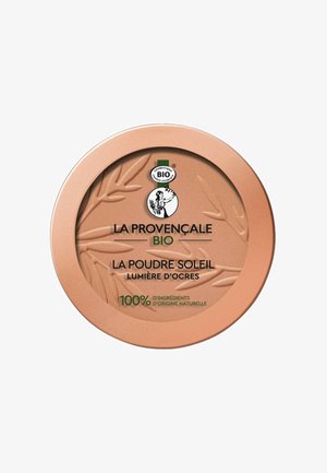 Compact rond en or rose avec un design de feuille texturé, comportant le nom du produit en noir et vert, ainsi qu'un texte indiquant des ingrédients naturels.