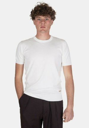 Giovane uomo con capelli ricci in piedi di fronte, indossa una camicia bianca attillata a maniche corte e pantaloni scuri, su uno sfondo chiaro e semplice.