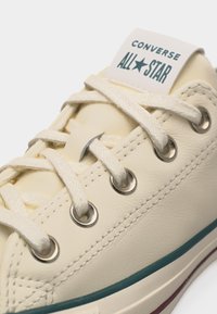 Παπούτσι Converse All Star σε κρεμ χρώμα με λευκά κορδόνια, μεταλλικά κλειδιά και πράσινη ρίγα στη σόλα.