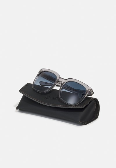 Óculos de sol grandes cinzentos com lentes escuras repousando sobre uma caixa de couro preto com costura castanha e um logótipo, também cosido.
