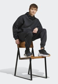 Black hooded sweatshirt and sweatpants, paired with black and white sneakers, seated on a wooden chair, featuring a minimalistic design.
