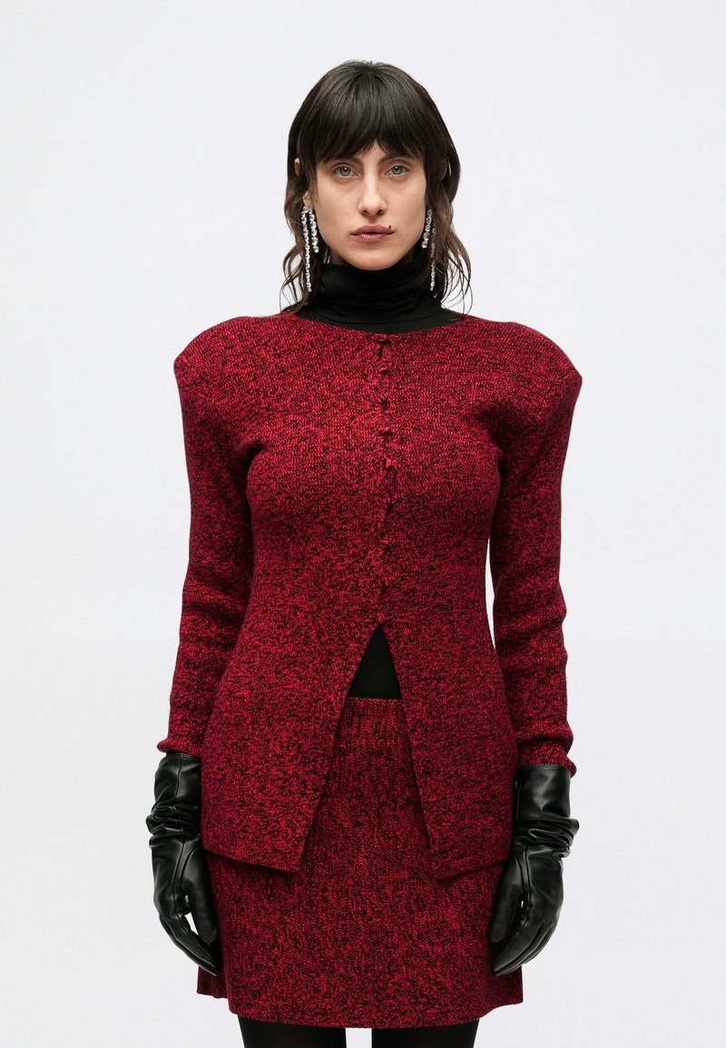 Červený textúrovaný pletený kabát s vypchávkami na ramenách, vertikálnym zapínaním na gombíky a vysokým rozparkom, v kombinácii so zladenou sukňou a čiernymi rukavicami.