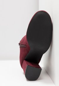 Bota de tobillo única de ante burdeos con tacón ancho, cremallera lateral y suela negra texturizada apoyada contra una pared blanca en esquina.