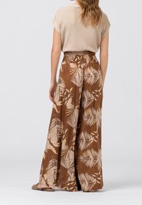 Hnědé květinové kalhoty s širokými nohavicemi, které mají smockovaný pas a vzory palmových listů. Vyrobené z lehké tkaniny s jemnou texturou.
