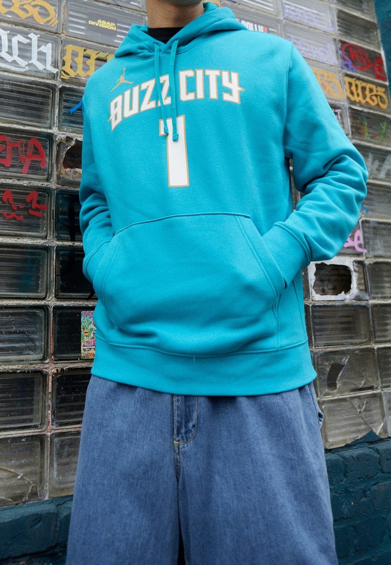 Felpa color teal con "BUZZ CITY" e un grande numero, caratterizzata da una tasca a marsupio e cappuccio con cordoncino, abbinata a pantaloni corti in denim blu.