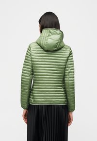 Γυναίκα που φοράει ανοιχτό πράσινο καπιτονέ μπουφάν με κουκούλα και μαύρη πλισέ φούστα, φωτογραφημένη από πίσω μπροστά σε απλό λευκό φόντο.