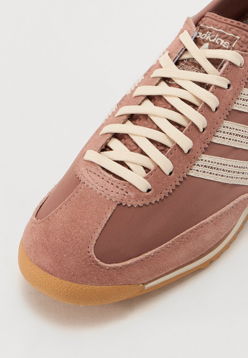 Sneaker marron présentant une combinaison de matériaux en daim et en tissu, avec des lacets crème, un design rayé et une semelle en caoutchouc texturée.