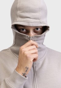 Hoodie zippé beige clair avec un col montant et une capuche. Comprend une fermeture éclair argentée et des poignets côtelés. Gros plan d'une main tenant la fermeture éclair.