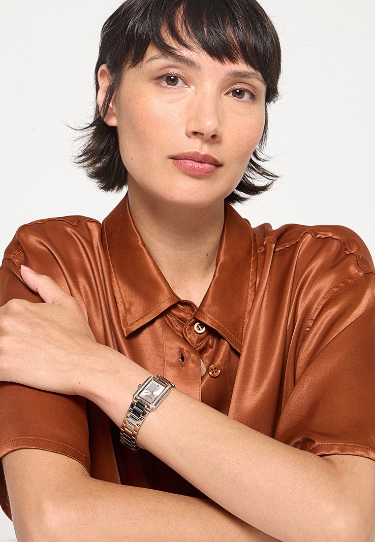Brązowa satynowa koszula z kołnierzykiem. Na lewym nadgarstku srebrno-złoty prostokątny zegarek z białą tarczą i metalową bransoletą.
