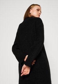 Lungo cappotto nero lavorato a maglia con una vestibilità rilassata, caratterizzato da maniche larghe e colletto alto. Il tessuto ha un aspetto morbido e testurizzato.