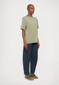Camiseta de manga corta en verde oliva con cuello redondo y un pequeño logo, combinada con unos vaqueros de mezclilla oscura y holgados, y zapatillas en color oliva.