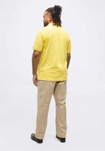 Άνδρας με ράστα μαλλιά, φορώντας κίτρινη πόλο μπλούζα, μπεζ παντελόνια και μαύρα παπούτσια, στέκεται με την πλάτη προς τα εμπρός σε λευκό απλό φόντο.