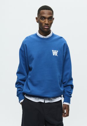 Kungligt blå sweatshirt med rund halsringning, sänkta axlar och en vit logotyp på vänster bröst, tillsammans med en vit skjorta med krage under.