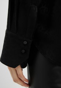 Manica di blusa nera con texture leopardata. Presenta due bottoni neri sul polsino. Tessuto liscio con una leggera lucentezza.