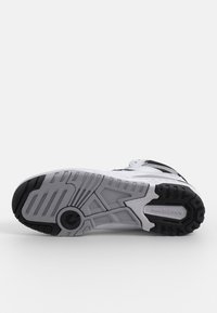 Vit sportskor med svarta detaljer, med en strukturerad gummisul, upphöjd mönster på yttersulan och en logotyp på hälen.