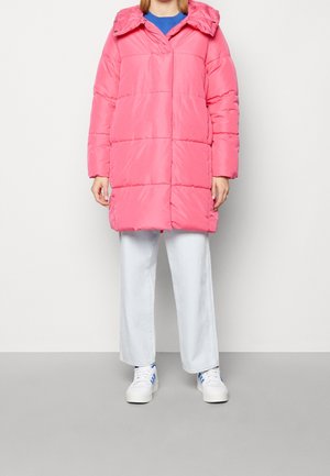 Personne portant un manteau matelassé rose vif mi-long, un pantalon blanc à jambes larges et des baskets blanches avec des rayures bleues sur fond uni.
