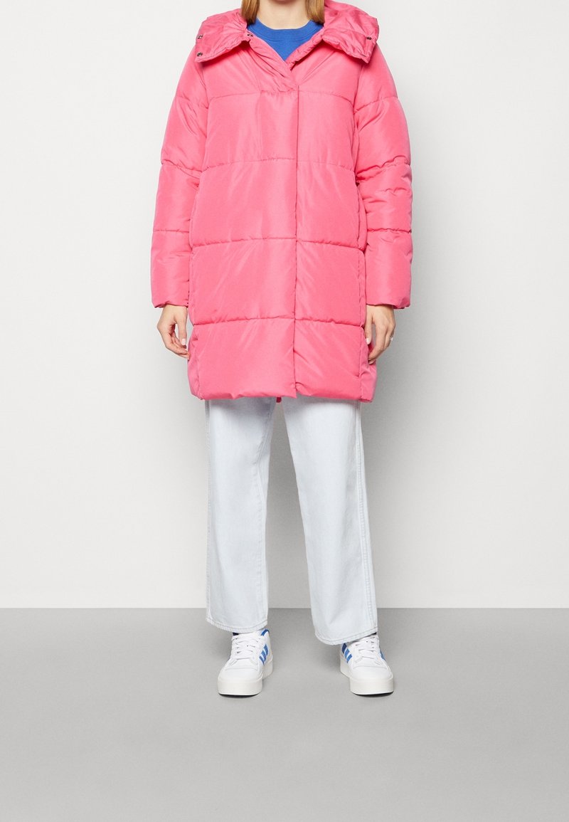 Personne portant un manteau matelassé rose vif mi-long, un pantalon blanc à jambes larges et des baskets blanches avec des rayures bleues sur fond uni.