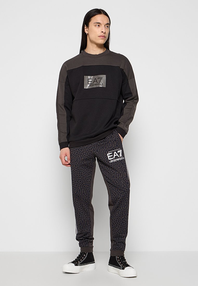 Felpa nera a maniche lunghe con dettagli grigi e un logo riflettente, abbinata a pantaloni della tuta neri caratterizzati da un motivo testurizzato e una branding bianca.