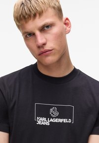 Juoda medvilninė marškinėliai trumpomis rankovėmis, su baltu logotipo langeliu, KL simboliu ir tekstu „KARL LAGERFELD JEANS“, atspausdintu priekyje.