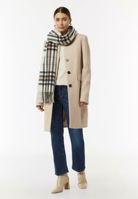 Lichtroze wollen jas met zwarte knopen, gelaagd over een crèmekleurige trui. Gecombineerd met donkerblauwe jeans en een geruite sjaal in beige, blauw en grijs.