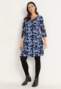 Blaues und schwarzes Kleid mit abstraktem Muster, drei Viertel Ärmel, ausgestellter Rock, kombiniert mit schwarzen Leggings und Stiefeletten.