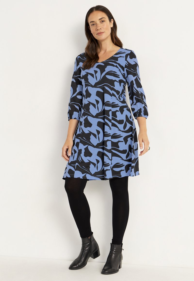 Blå og sort abstrakt-mønstret kjole med trekvartærmer, flagrende skirt, og kombineret med sorte leggings og ankelstøvler.