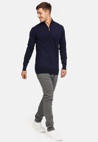 Pull-over long de couleur marine avec un col zippé, associé à un pantalon slim gris clair et des baskets blanches. Présente une texture lisse.