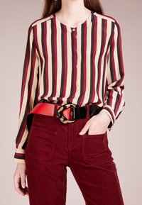 Blouse rayée en beige, rouge et noir ; fermeture à boutons. À associer avec un pantalon en velours côtelé bordeaux et une large ceinture noire avec une boucle à motif.