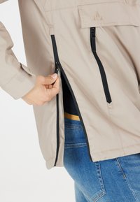 Beige jacka med svarta dragkedjor, med sidofickor och en strukturerad yta. Modellen visar dragkedjedetaljer mot denim.