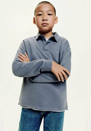 Grijze polo met lange mouwen en een kraag, met zijsplitten, gedragen over een blauwe spijkerbroek. De stof lijkt zacht en heeft een casual pasvorm.