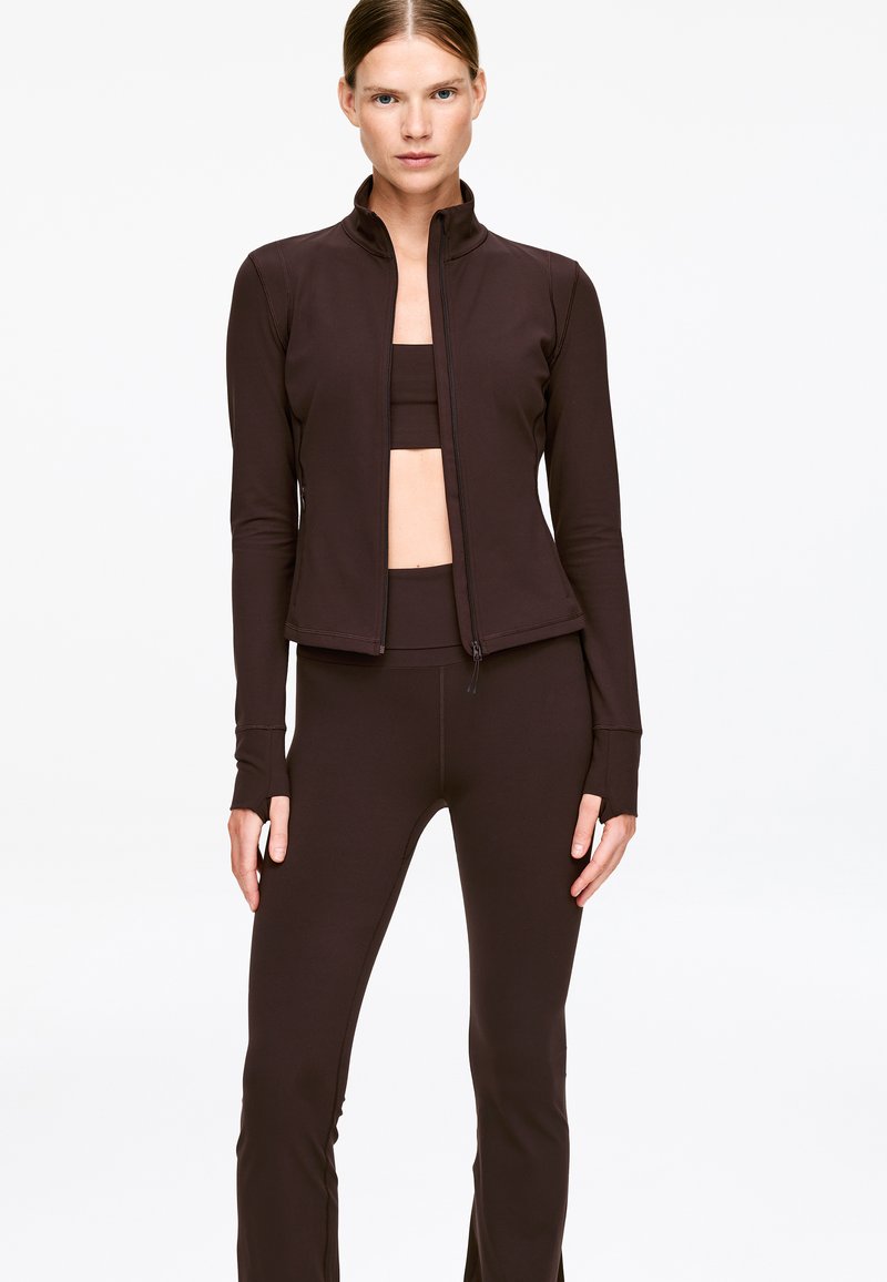 Veste de sport marron avec col haut et fermeture éclair devant, associée à un top court assorti et un pantalon ajusté. Tissu lisse, silhouette élégante.