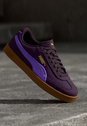 Violetti Puma-tennari, jossa laventelivärinen raita ja kumipohja, näkyy leijumassa teksturoidun harmaan pinnan yläpuolella.