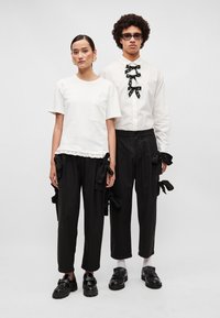 T-shirt branca de algodão com bolso e bainha com babados; calças pretas com laços nas laterais. Modelo usa sapatos pretos; segundo modelo em camisa branca com detalhe em laço.