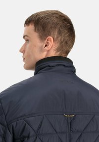 Marineblå quiltet jakke med høj krave og tekstureret syning. Har et logo på ryggen og en lille dekorativ detalje på kraven.