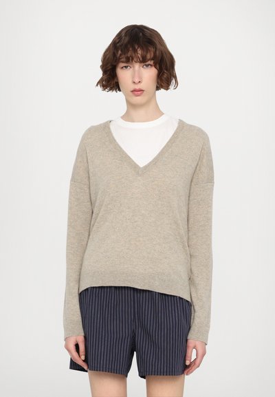 Pull en tricot beige avec un col en V, des manches longues et une coupe décontractée. Porté sur un t-shirt blanc et associé à un short rayé bleu marine.