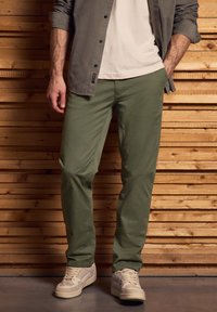 Vihreät puuvillahousut slim fit -mallia, yhdistettynä vaaleisiin lenkkareihin ja harmaaseen napitettuun paitaan, joka on kerrostettu valkoisen T-paidan päälle.