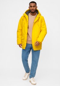 Man in een gele winterjack met capuchon, beige sweater, blauwe spijkerbroek en witte sneakers, staande tegen een egale witte achtergrond.