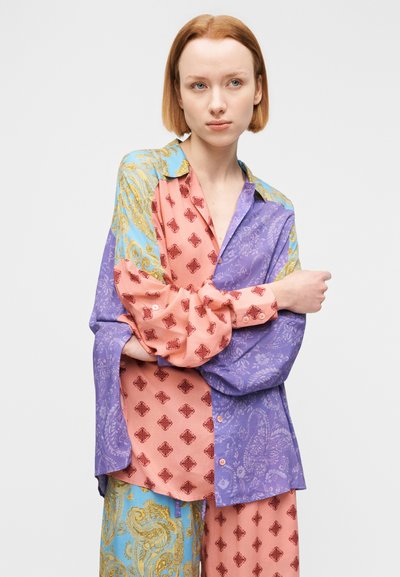 Mladá osoba s krátkymi červenými vlasmi, oblečená v voľnej viacfarebnej vzorovanej blúzke a zladených nohaviciach, stojaca pred jednoduchým pozadím.