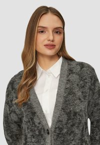 Grijze cardigan met een gestructureerde afwerking, open voorkant en een enkele knop. Draagt over een witte collared shirt. Zachte stof met een subtiele glans.