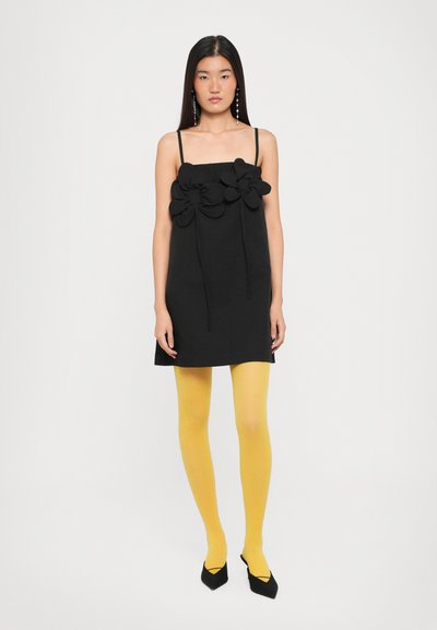 Fekete, ujjatlan ruha, himbálózó virágdíszítésekkel, élénksárga harisnyával és fekete, hegyes orrú magassarkú cipővel párosítva.