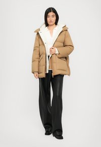Teddy-Jacke in Beige mit einer weißen, mit Fell gefütterten Kapuze. Sie hat einen Reißverschluss, eine horizontale Steppung und wird mit schwarzen Lederhosen kombiniert.