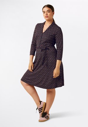 Vestido envolvente de tela oscura con un estampado floral, mangas tres cuartos y un cinturón. Combinado con zapatillas rosas y cordones negros.