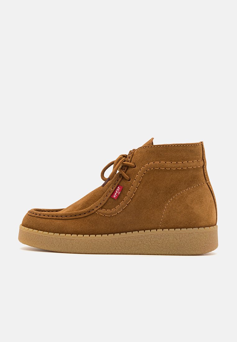 Bottines en suède beige avec un bout arrondi, des lacets, une semelle en caoutchouc beige et des coutures décoratives. Étiquette logo rouge sur le côté.
