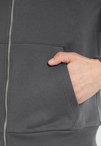 Hoodie zippé gris en mélange de coton doux, avec une poche avant, un ourlet et des poignets côtelés, et des détails de couture visibles le long des coutures.