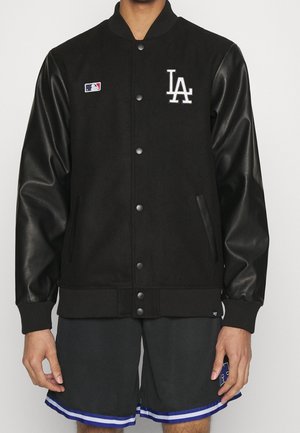 Person iført en sort varsity-jakke med læderærmer og LA-logo på brystet, kombineret med sorte og blå atletikshorts.