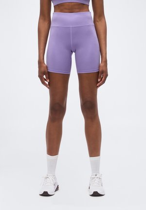 Person iført lilla cykelshorts med høj talje, hvide knæstrømper og hvide sportssko, står mod en ensartet lys baggrund.
