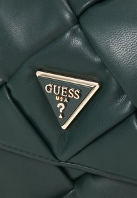 Sac à main matelassé vert avec une texture lisse, présentant une plaque en forme de triangle doré. Le design comprend des accents rembourrés et une finition brillante.